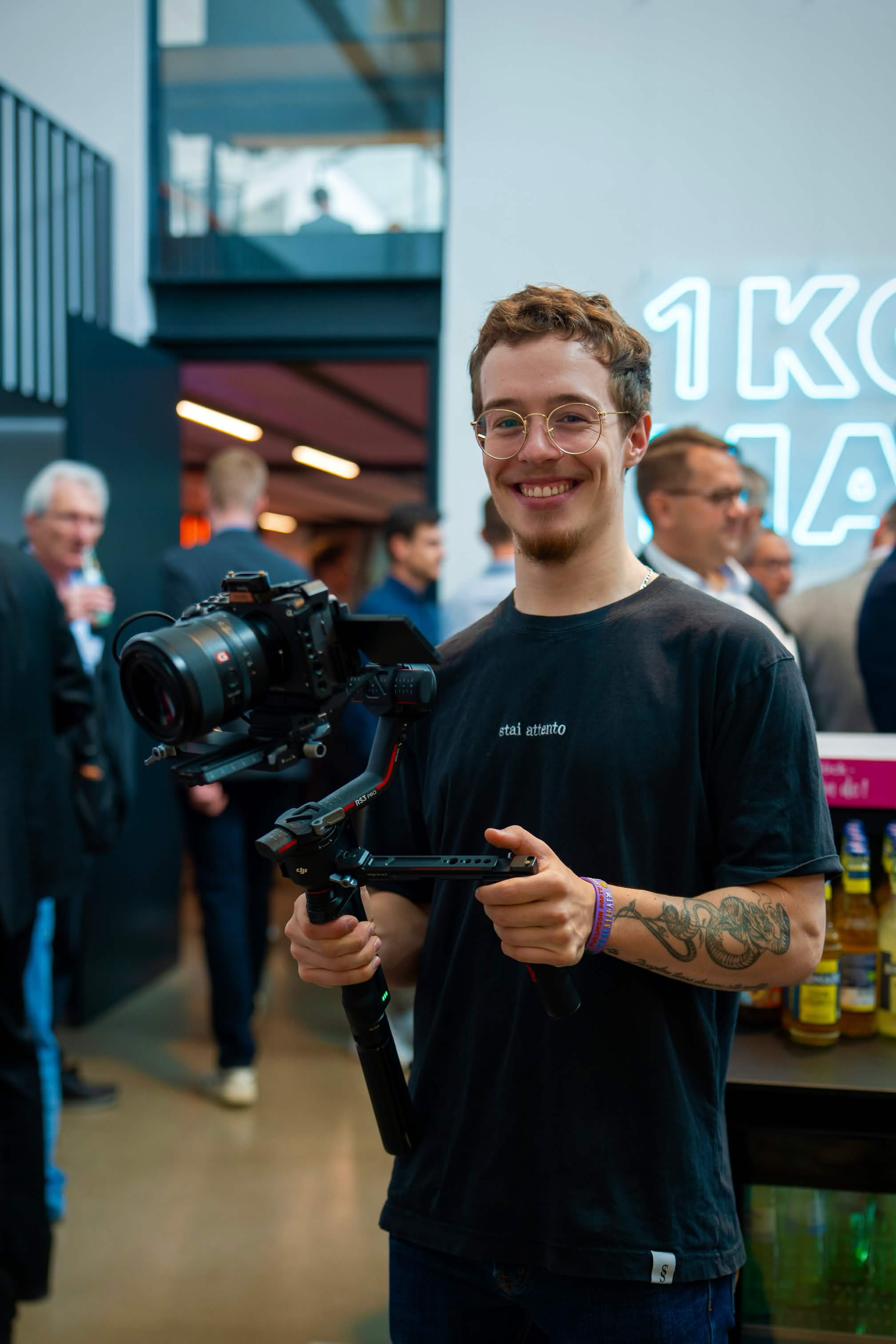 Tobiasz Smith smiling at the camera while holding a professional video camera on a gimbal stabilizer. He wears glasses and a black t-shirt with a tattoo visible on his arm. Behind him is a modern venue with blue architectural elements, neon signage, and other people in business attire. The atmosphere is professional and welcoming.